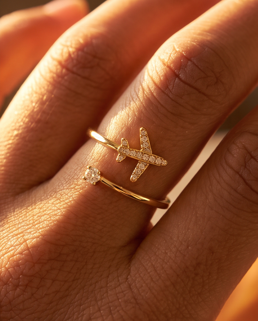 A close-up of a hand wearing an adjustable gold plated ring with a small airplane charm paved with tiny clear stones set against warm golden light.
