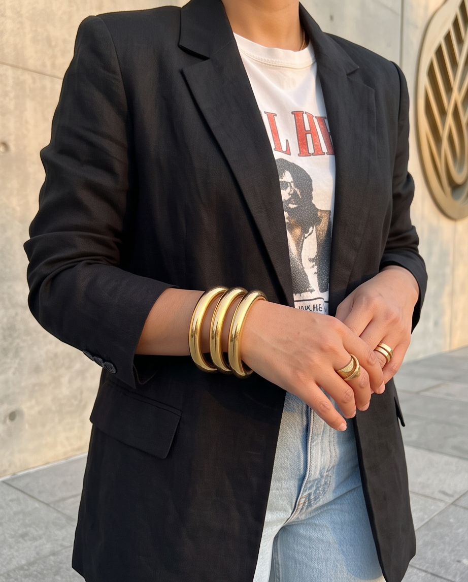 A person wearing a black blazer over a white graphic T-shirt and light blue jeans, with multiple gold bangles and rings on the hands, standing outdoors against a concrete wall.