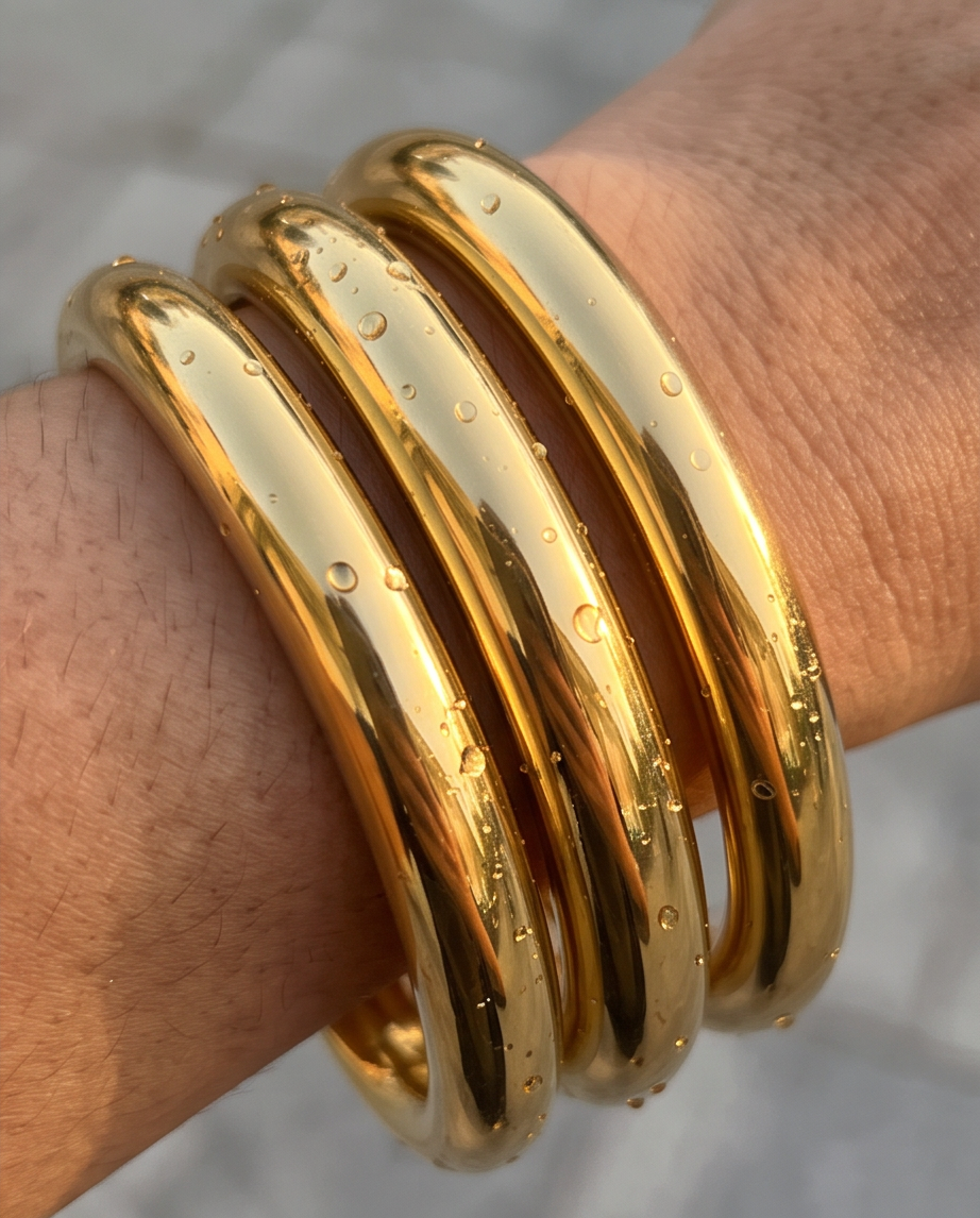 A close-up of a person's wrist wearing a stack of 3 chunky gold polished bangles, with water droplets on their surface, tarnish resistan bangles, waterproof and hypoallergenic stainless steel bangles for woman. 