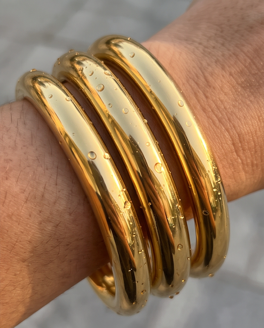 A close-up of a person's wrist wearing a stack of 3 chunky gold polished bangles, with water droplets on their surface, tarnish resistan bangles, waterproof and hypoallergenic stainless steel bangles for woman. 