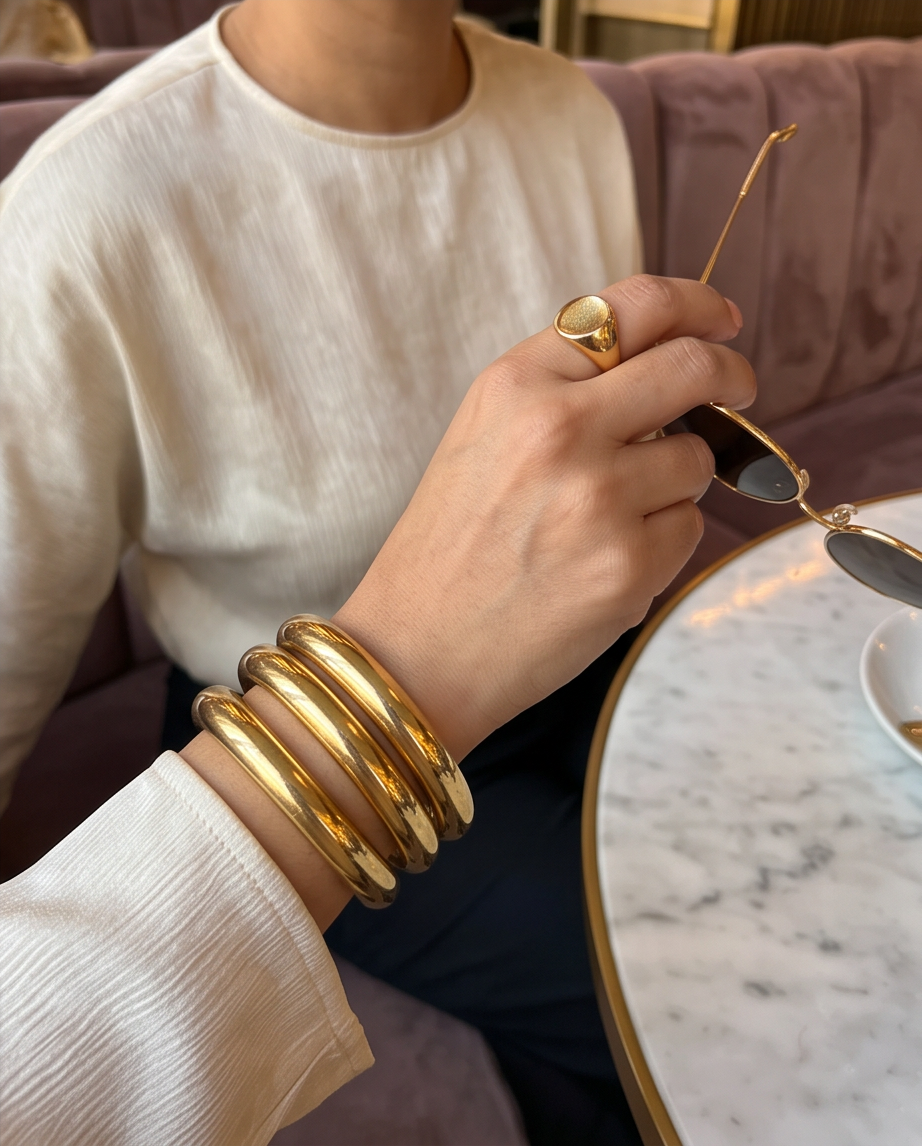 A close-up lifestyle shot of a person wearing a stack of three polished gold bangle bracelets on their wrist, holding a pair of gold-framed sunglasses. The person is wearing a cream-colored textured top.