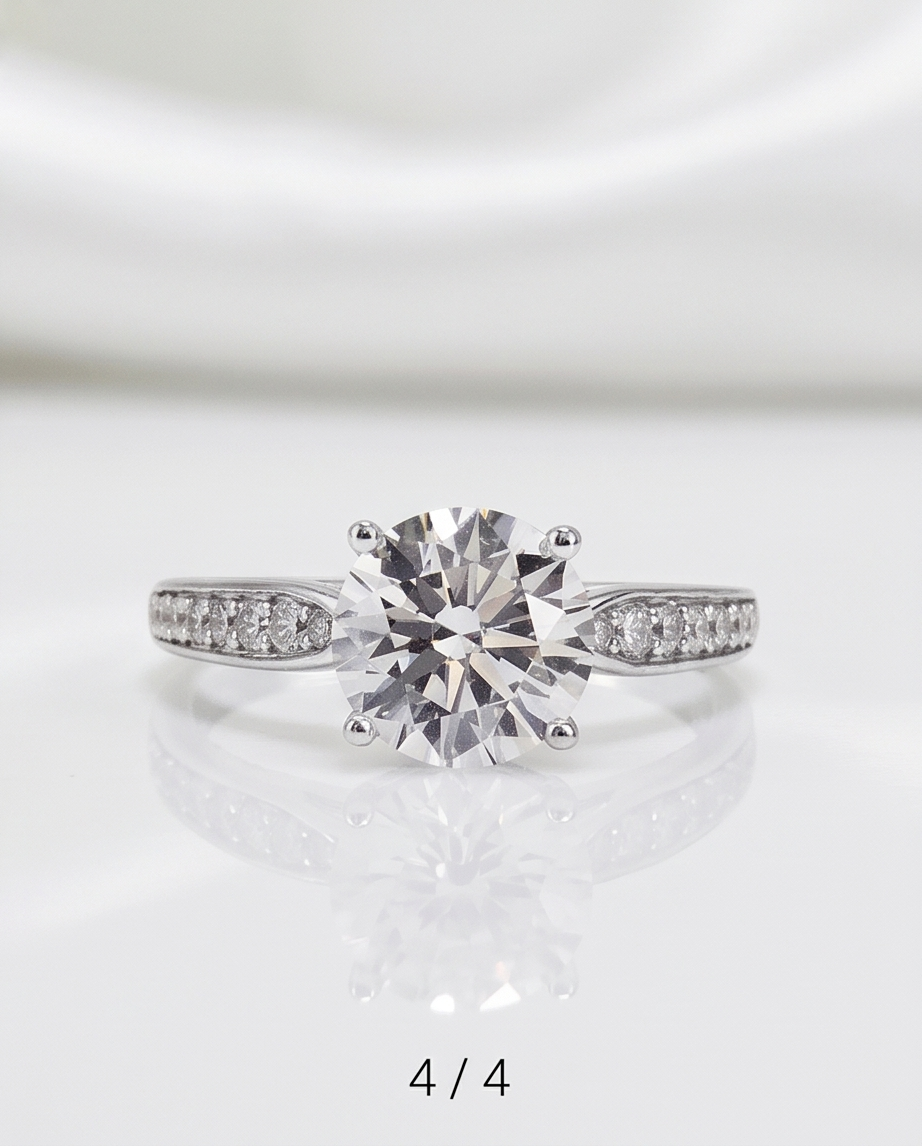 A round-cut solitaire engagement ring with a silver-toned band set with small pavé diamonds, displayed on a reflective white surface with a soft satin background.