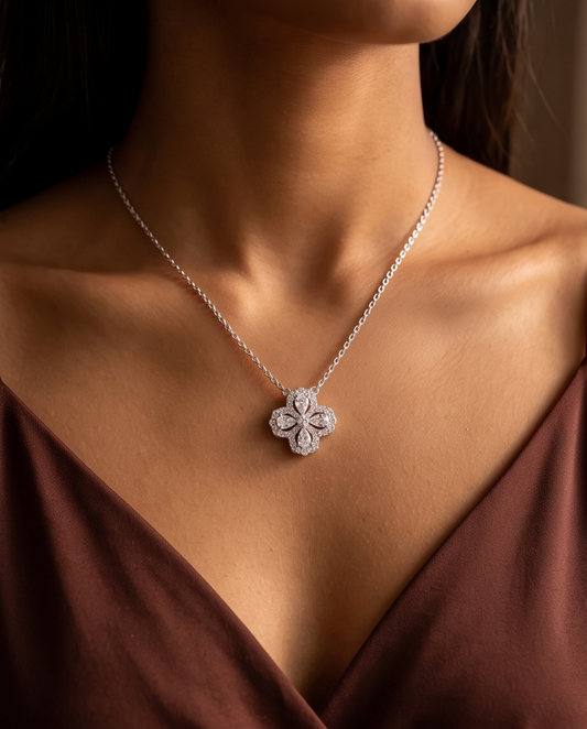 A close-up of a model wearing a delicate silver chain necklace with a four-petal flower pendant paved with small clear stones, styled with a dark brown satin top.
