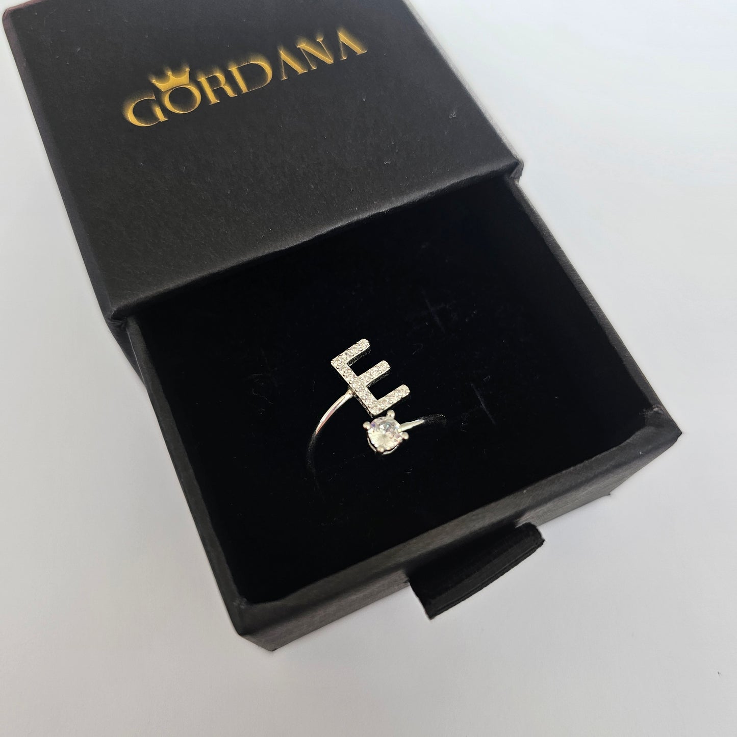 Silver alphabet ring with letter E and a small gemstone, stainless steel fashion jewelry in a black Gordana box.