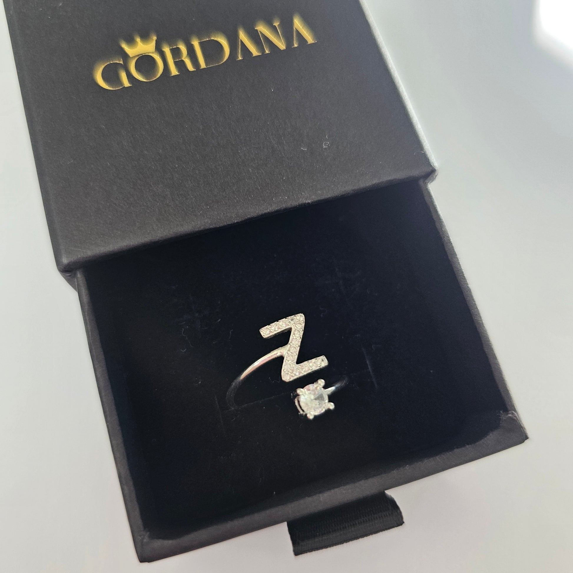 Silver alphabet ring with gemstone detail in black velvet box, stainless steel jewelry, unisex fashion style.