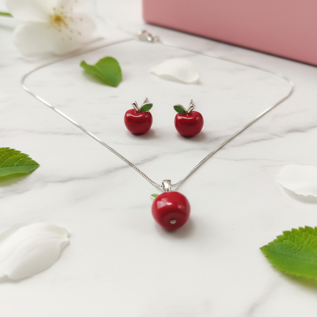 Red apple-shaped necklace and earrings on a white surface with green leaves and a pink box. Silver Jewelry set