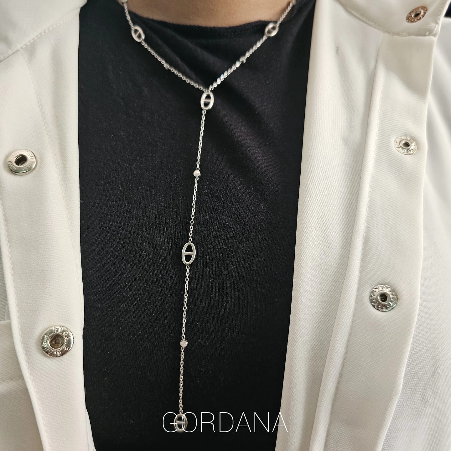 Silver stainless steel necklace with elongated chain links and circular accents, worn over black shirt and white jacket.