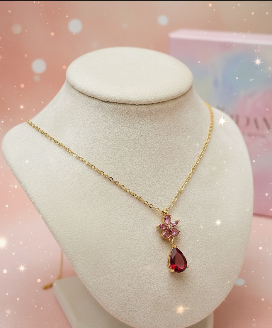 Gold necklace with a red teardrop pendant on a white stand against a pink background