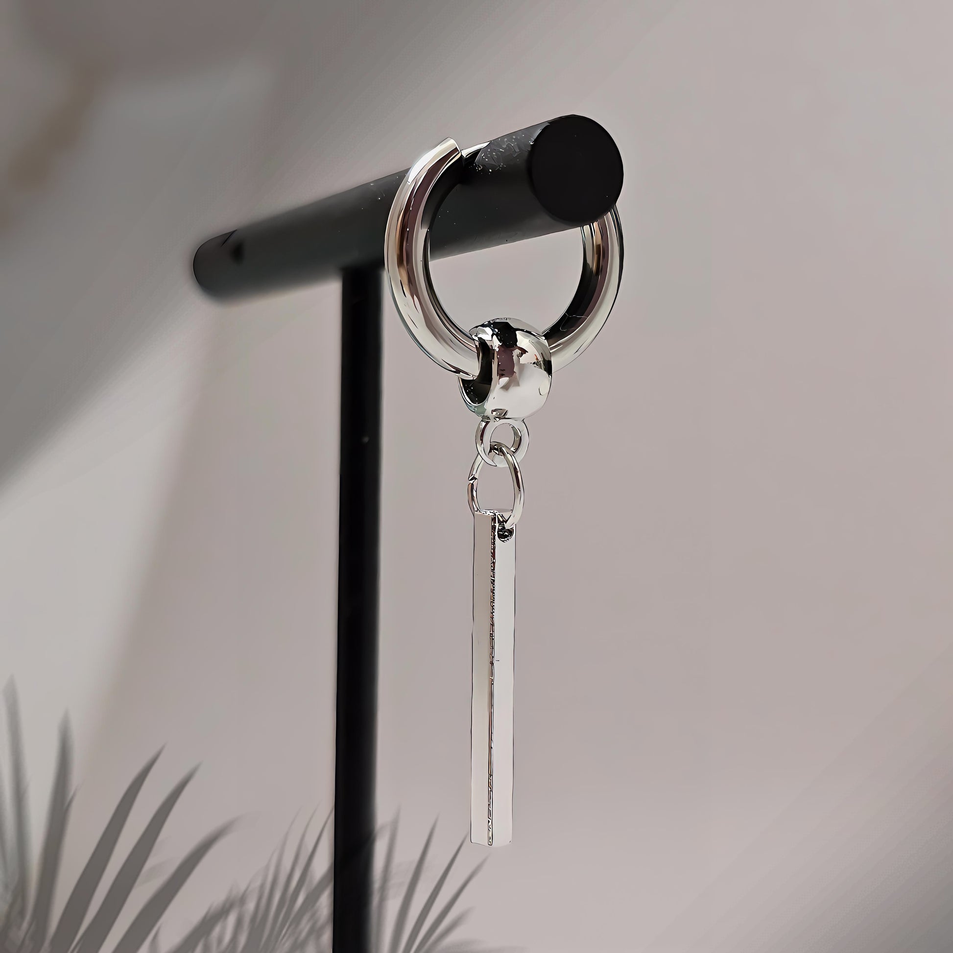 Silver earring with a dangling bar on a black stand against a neutral background