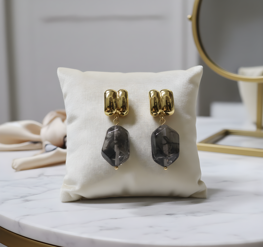 Gold and gray smokey pendant earrings on a white cushion with a blurred background