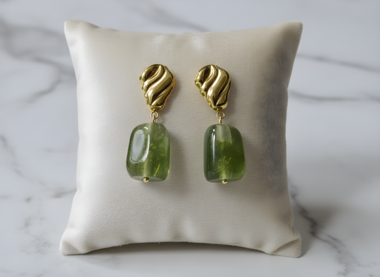 Gold earrings with green gemstones on a beige pillow against a marble background