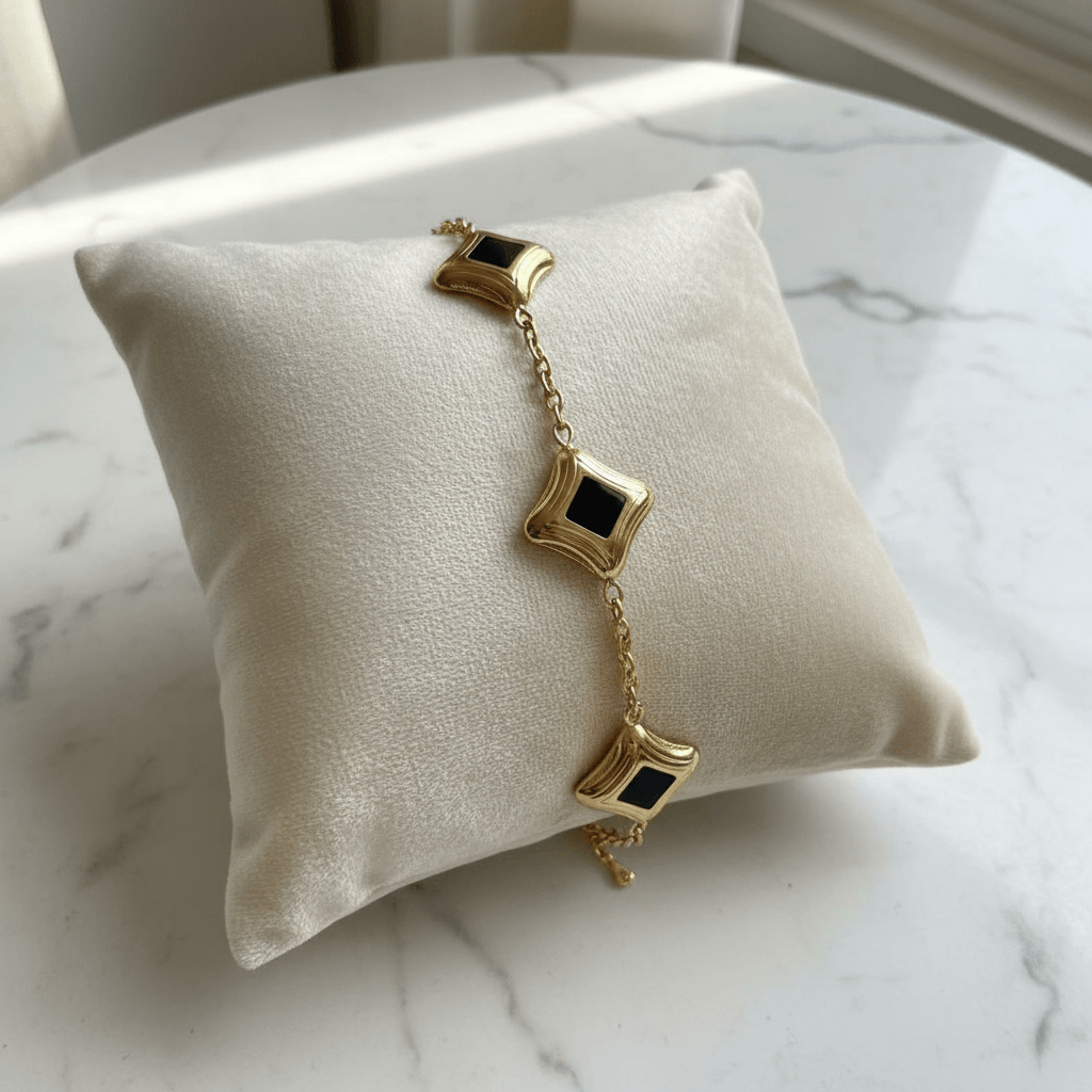 Gold bracelet with black square charms on a white cushion against a marble background