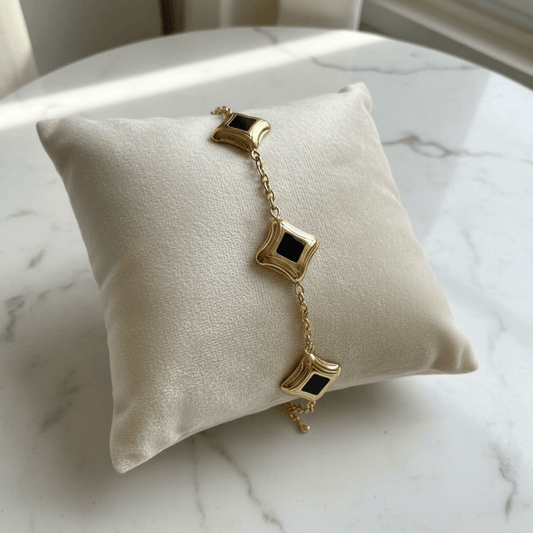 Gold bracelet with black square charms on a white cushion against a marble background